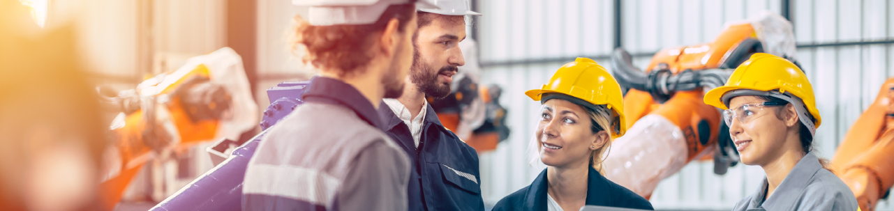 Four employees in hard hats in conversation on the floor of a manufacturing facility